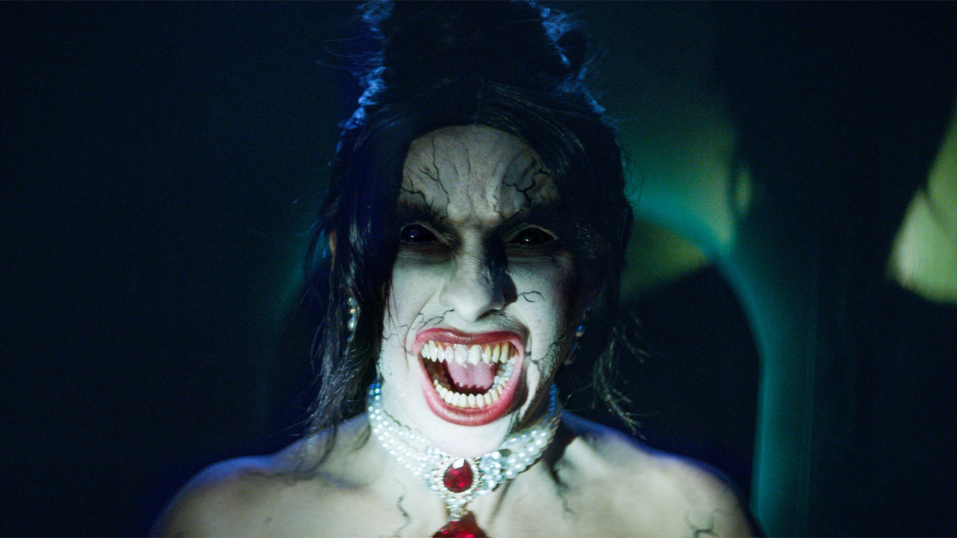 A person in heavy horror makeup snarls, showing sharp fangs, cracked white face paint, dramatic dark eye makeup, a jeweled necklace, and dark hair, illuminated by eerie, dim lighting.