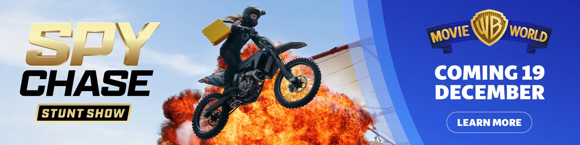 A motorcyclist in black gear jumps in front of a fiery explosion. Text reads "Spy Chase Stunt Show" and "Movie World Coming 19 December." A blue button says "Learn More.