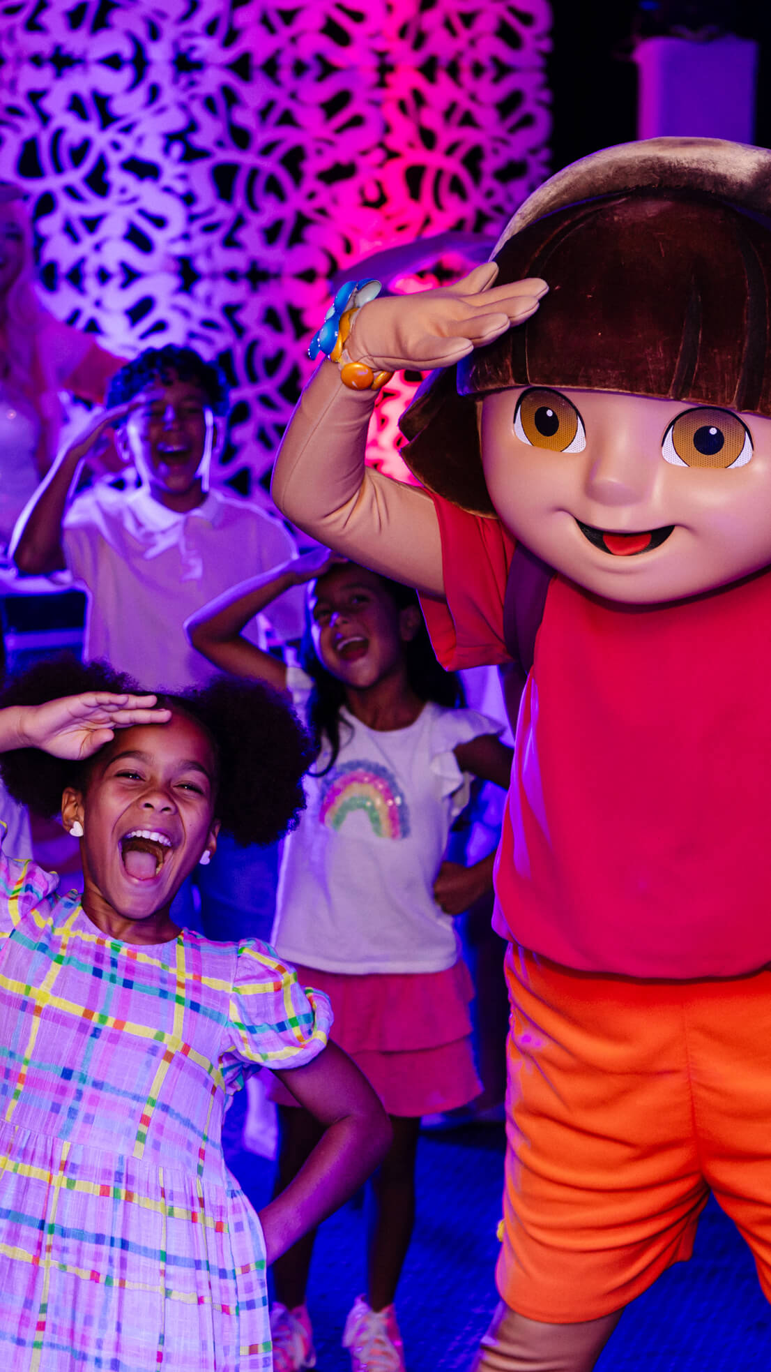 A child in a plaid dress and others in the background pose with Dora the Explorer at an event with colorful lighting.