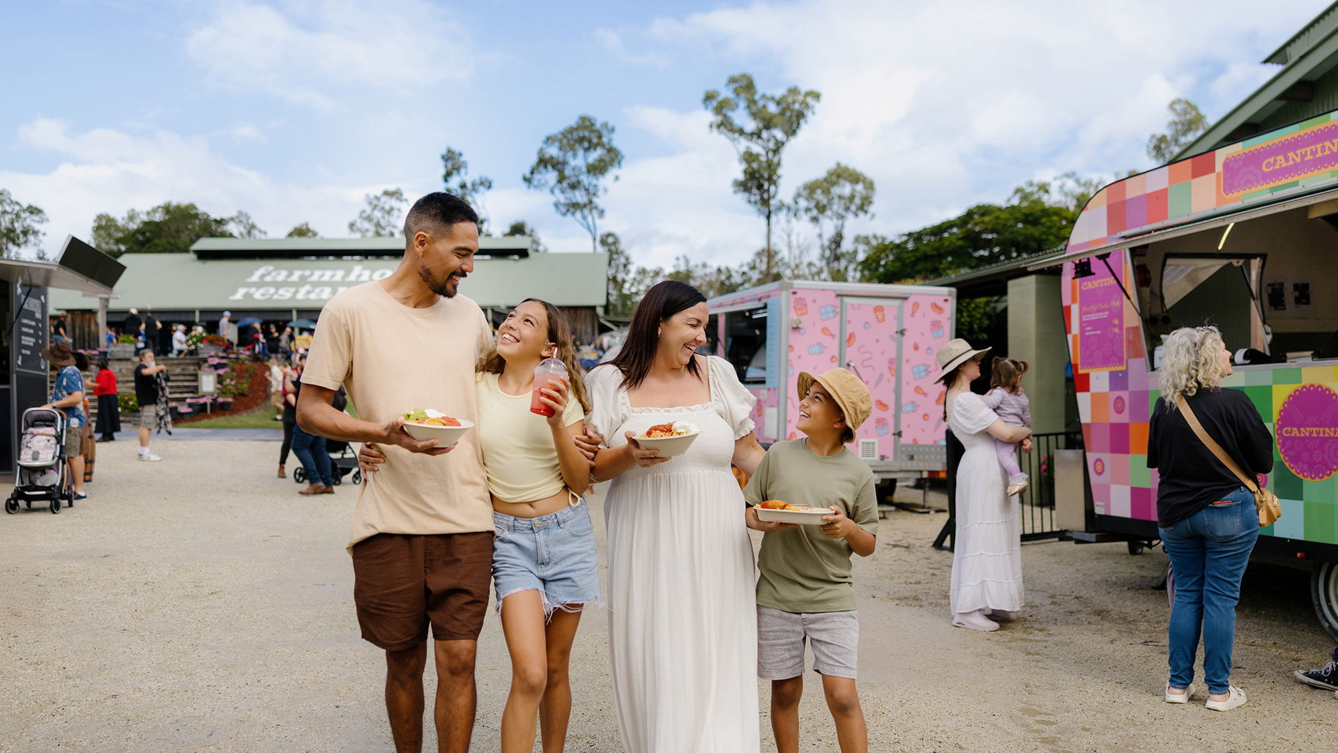 A family of four, smiling and holding food, walks together at an outdoor food market with colorful food trucks and people in the background on a sunny day.