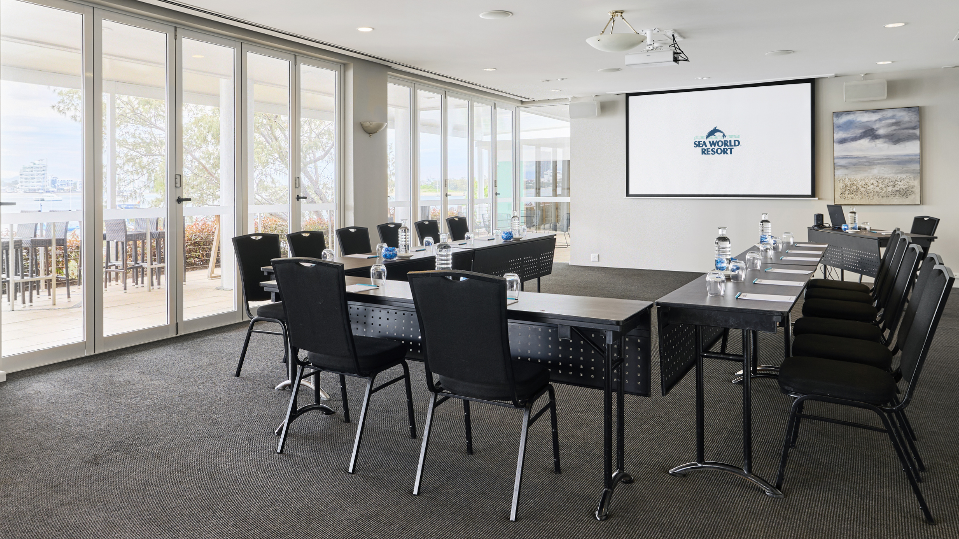 Modern conference room with U-shaped tables, black chairs, water bottles, notepads, and pens. Large windows let in natural light, and a projector screen displays “The Ville Resort.” Outdoor area visible through glass doors.
