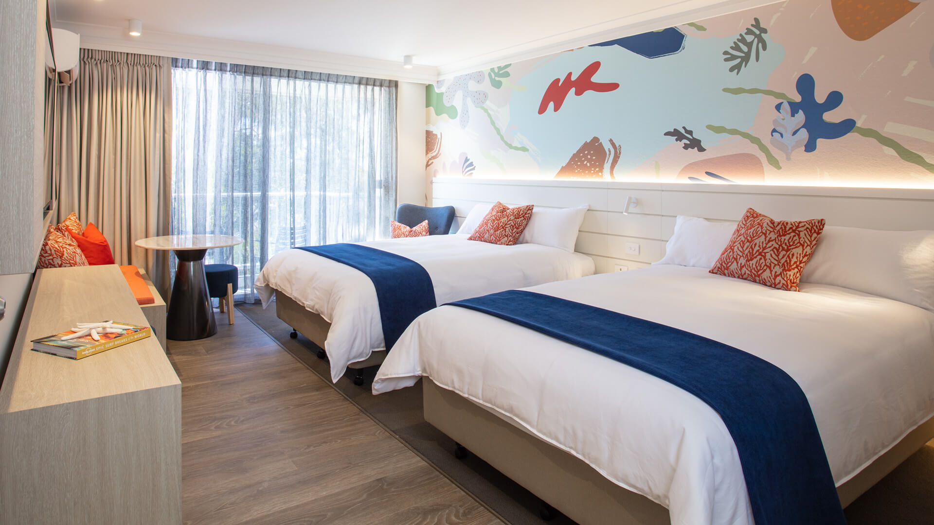 Bright hotel room with two double beds, blue accents, decorative pillows, a patterned mural wall, floor-to-ceiling window with sheer curtains, and a small table with chairs.
