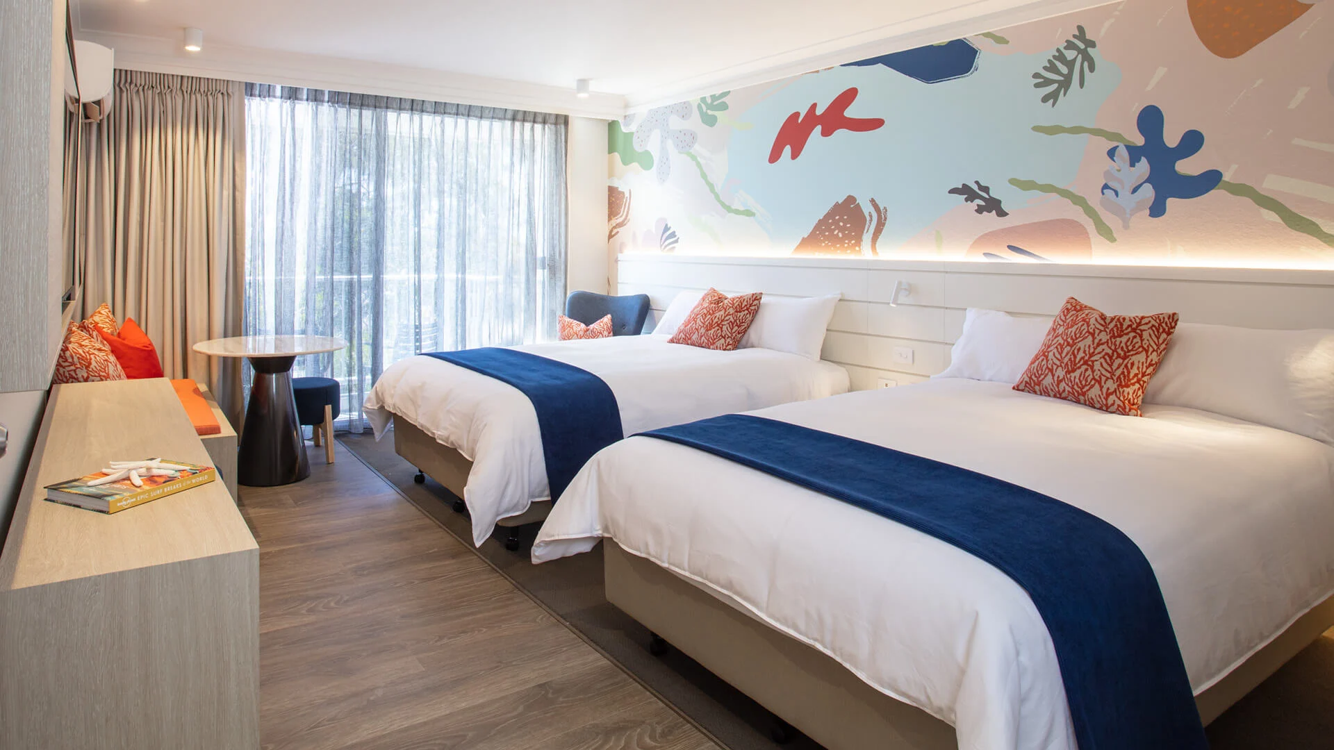 Bright hotel room with two double beds, blue accents, decorative pillows, a patterned mural wall, floor-to-ceiling window with sheer curtains, and a small table with chairs.