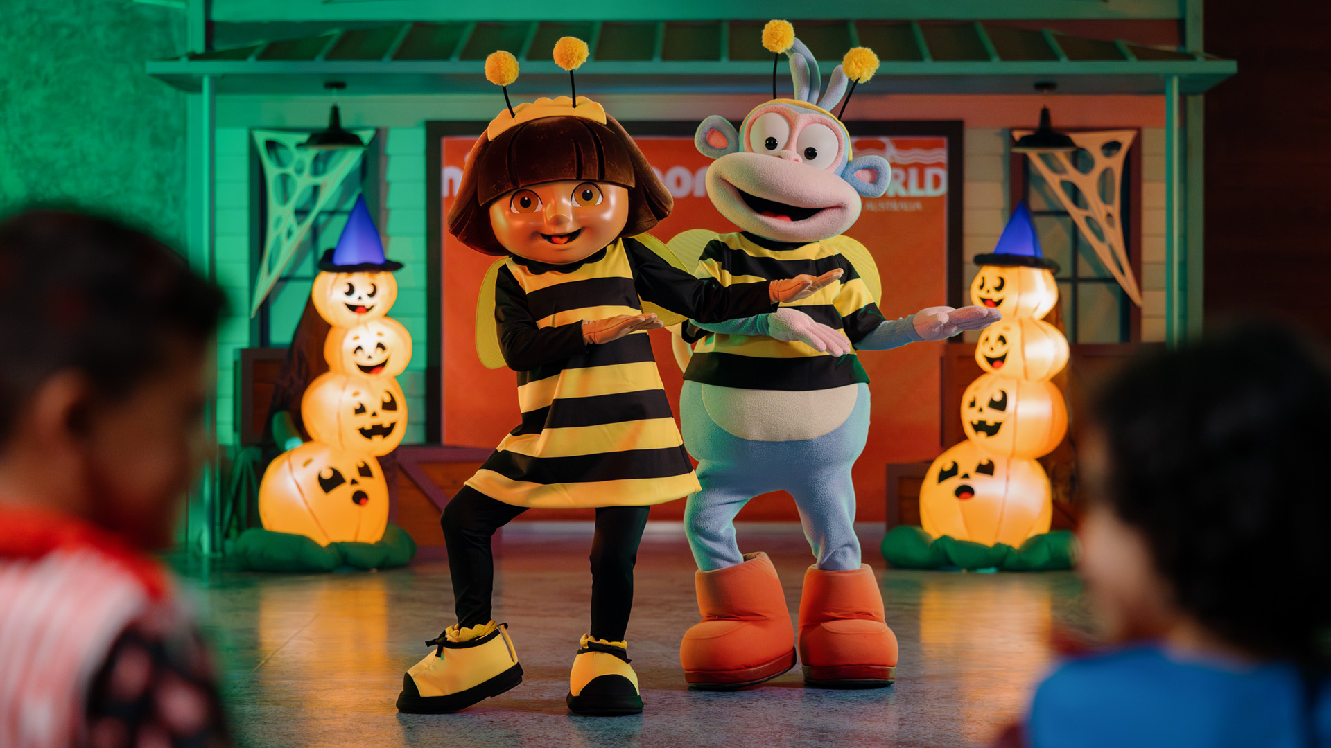 Two costumed characters, one dressed as a bee and the other as a blue and white monkey, dance in front of glowing Halloween pumpkin decorations while children watch. The scene is festive and colorful.