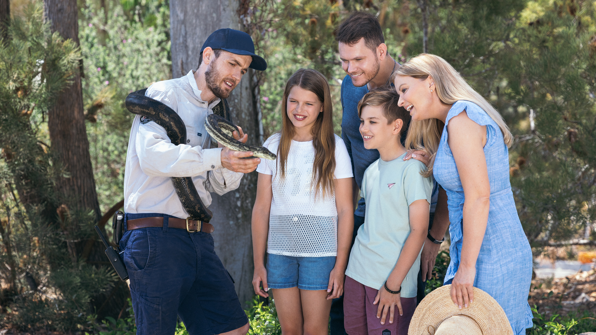 A man holding a large snake shows it to a smiling family of four, including two children, outdoors with trees in the background.