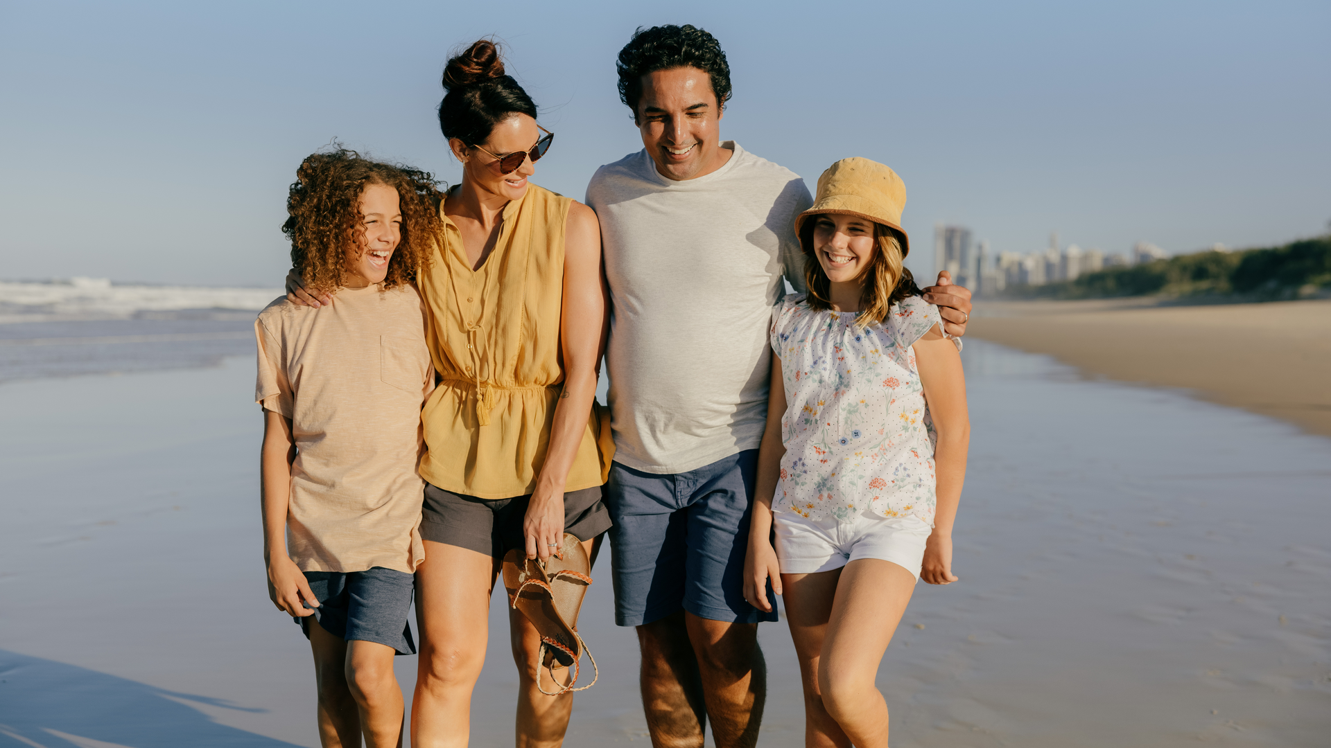 A family of four walks along a sandy beach, smiling and enjoying the sunny weather. The parents have their arms around their two children. Ocean waves and city buildings are visible in the background.
