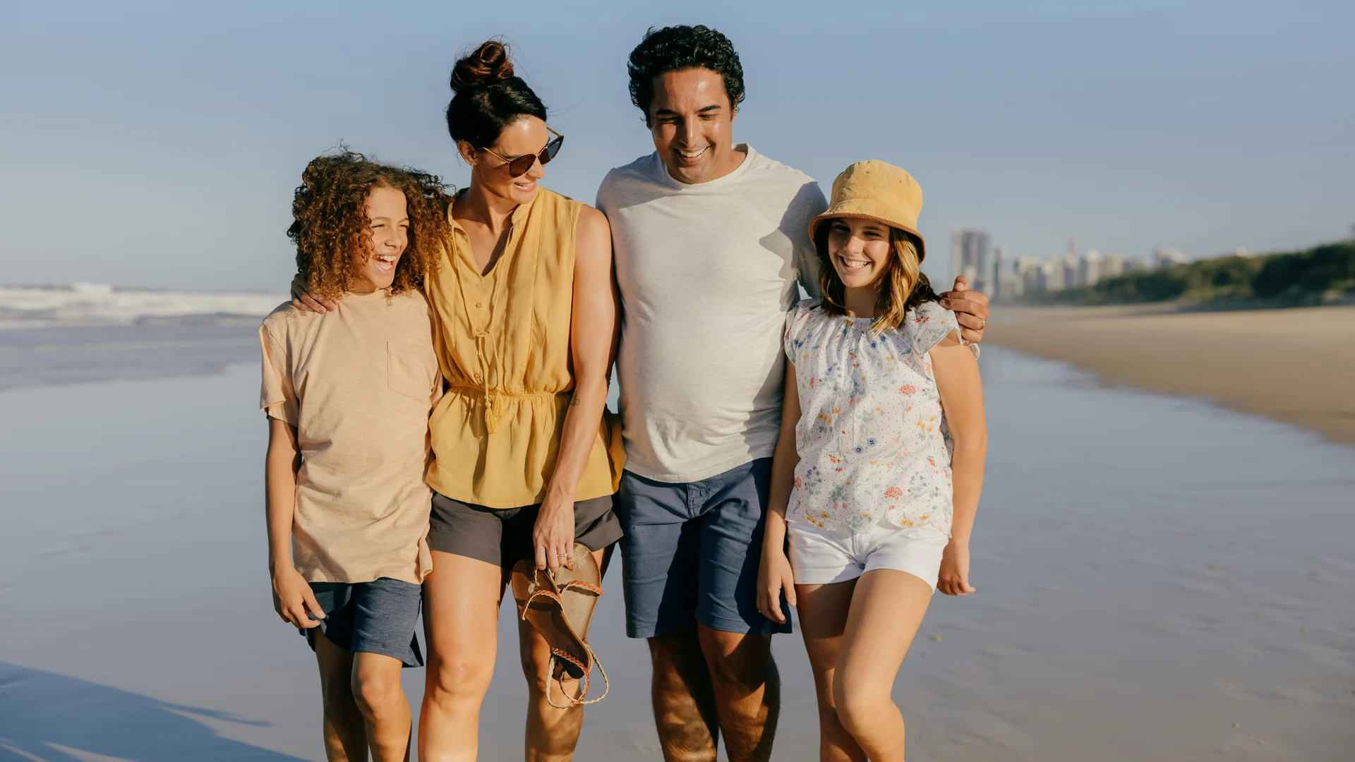 A family of four walks along a sandy beach, smiling and enjoying the sunny weather. The parents have their arms around their two children. Ocean waves and city buildings are visible in the background.