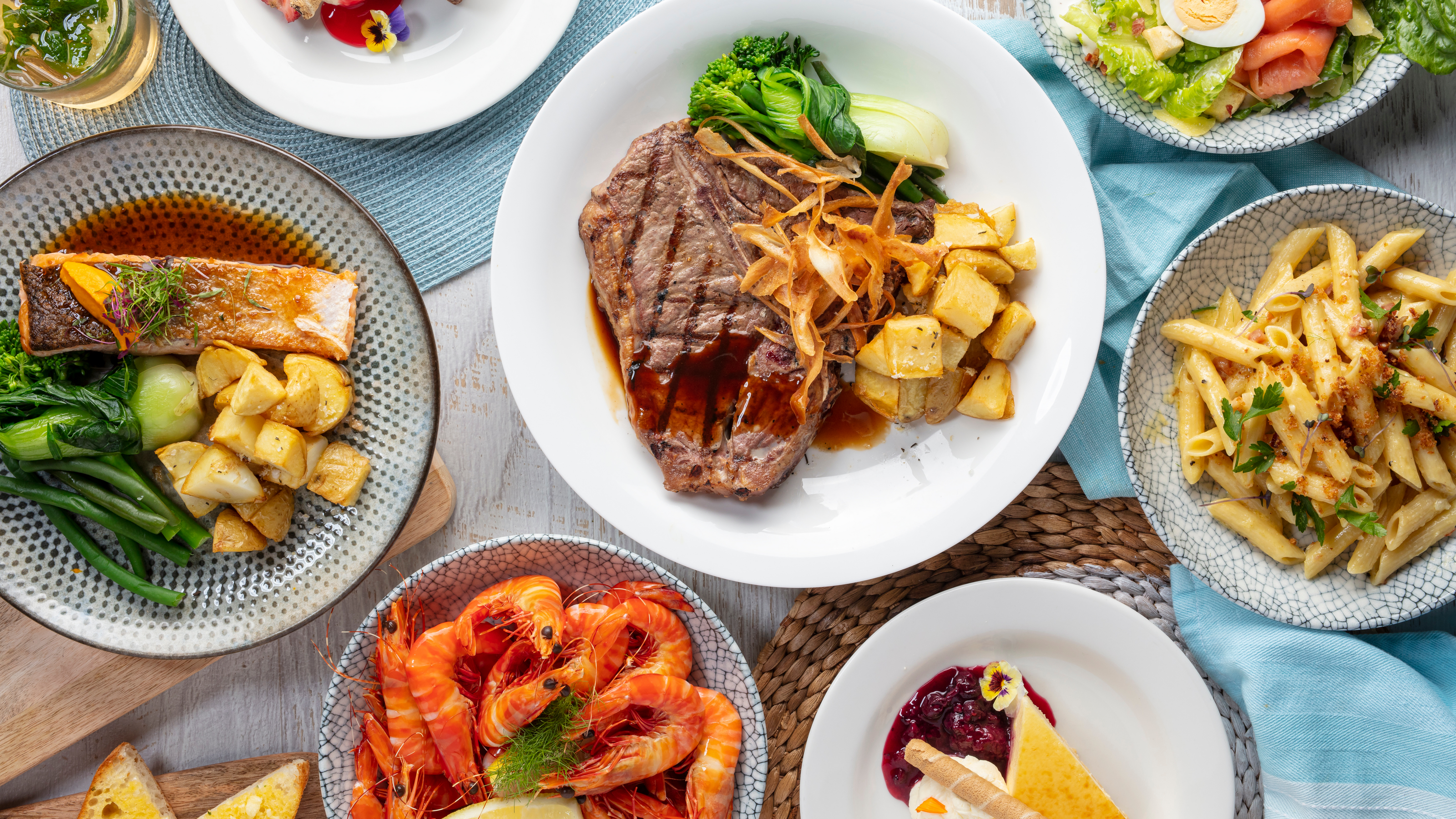 A table with various dishes, including steak, salmon, prawns, fries, a vegetable salad, and dessert with a slice of cake and berry sauce.