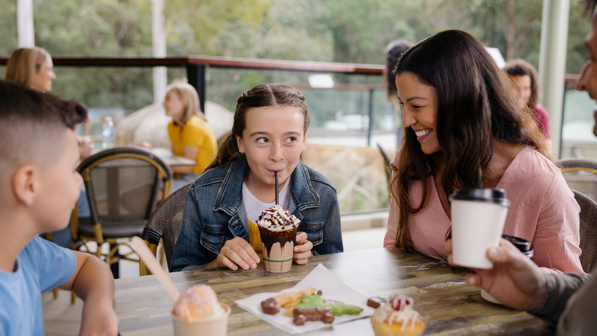 A girl drinks a milkshake with whipped cream and chocolate drizzle, sitting at a table with a woman and children, surrounded by desserts and drinks in a café setting.
