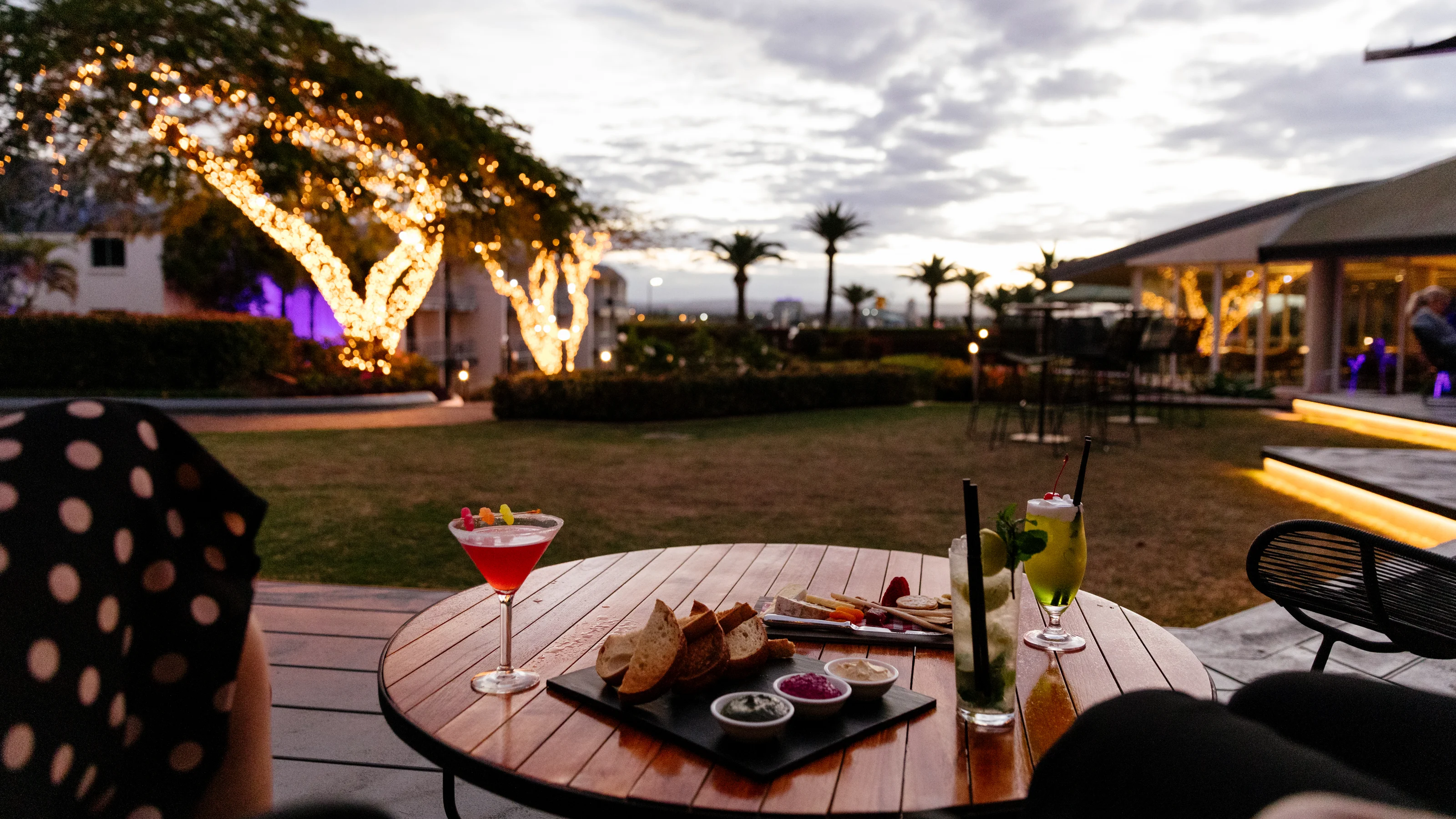 Outdoor seating area on the Lobby Deck at dusk with a wooden table holding cocktails, bread, and dips. Trees are decorated with lights, and a cloudy sky is in the background.