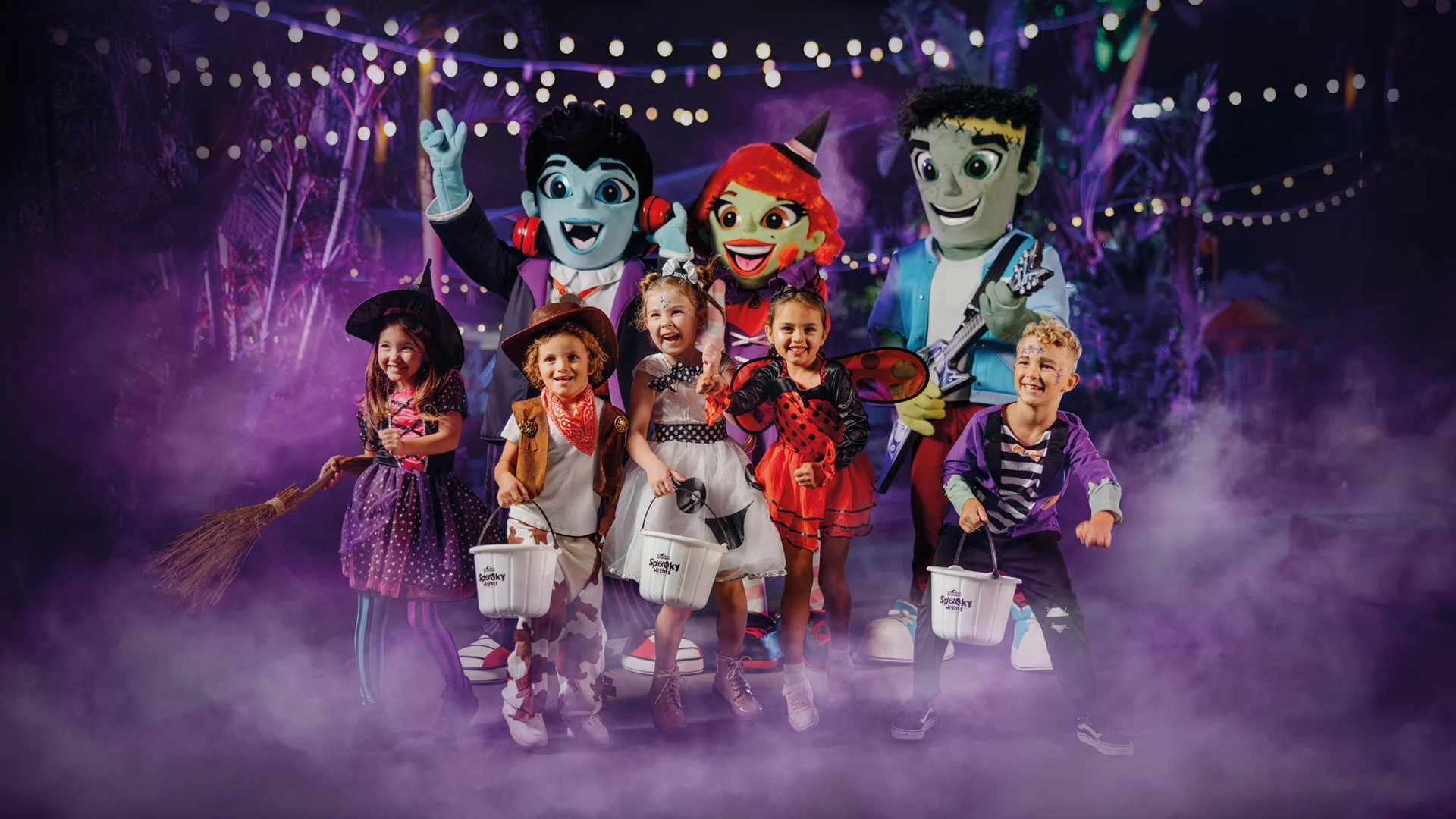 A group of young children in Halloween costumes stand smiling with trick-or-treat buckets, surrounded by friendly cartoon monster mascots, under string lights and purple mist in a festive nighttime setting.