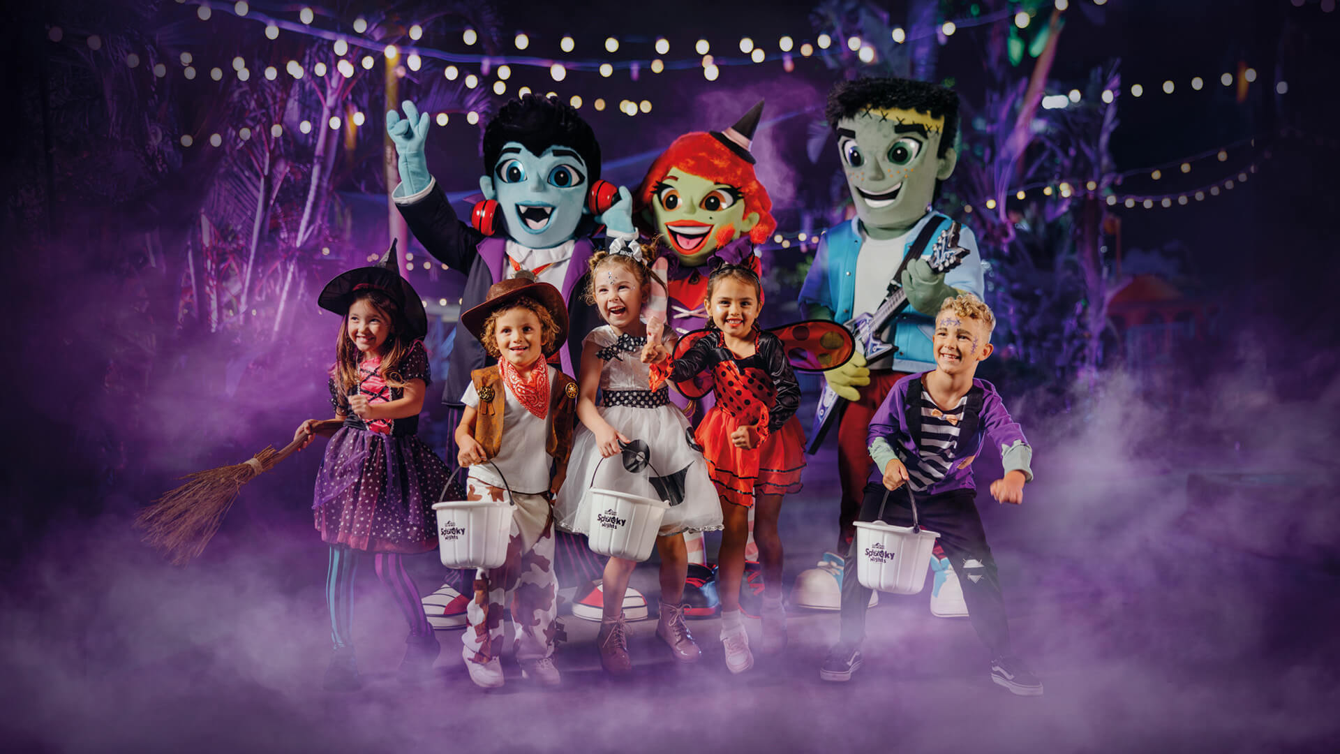 A group of young children in Halloween costumes stand smiling with trick-or-treat buckets, surrounded by friendly cartoon monster mascots, under string lights and purple mist in a festive nighttime setting.