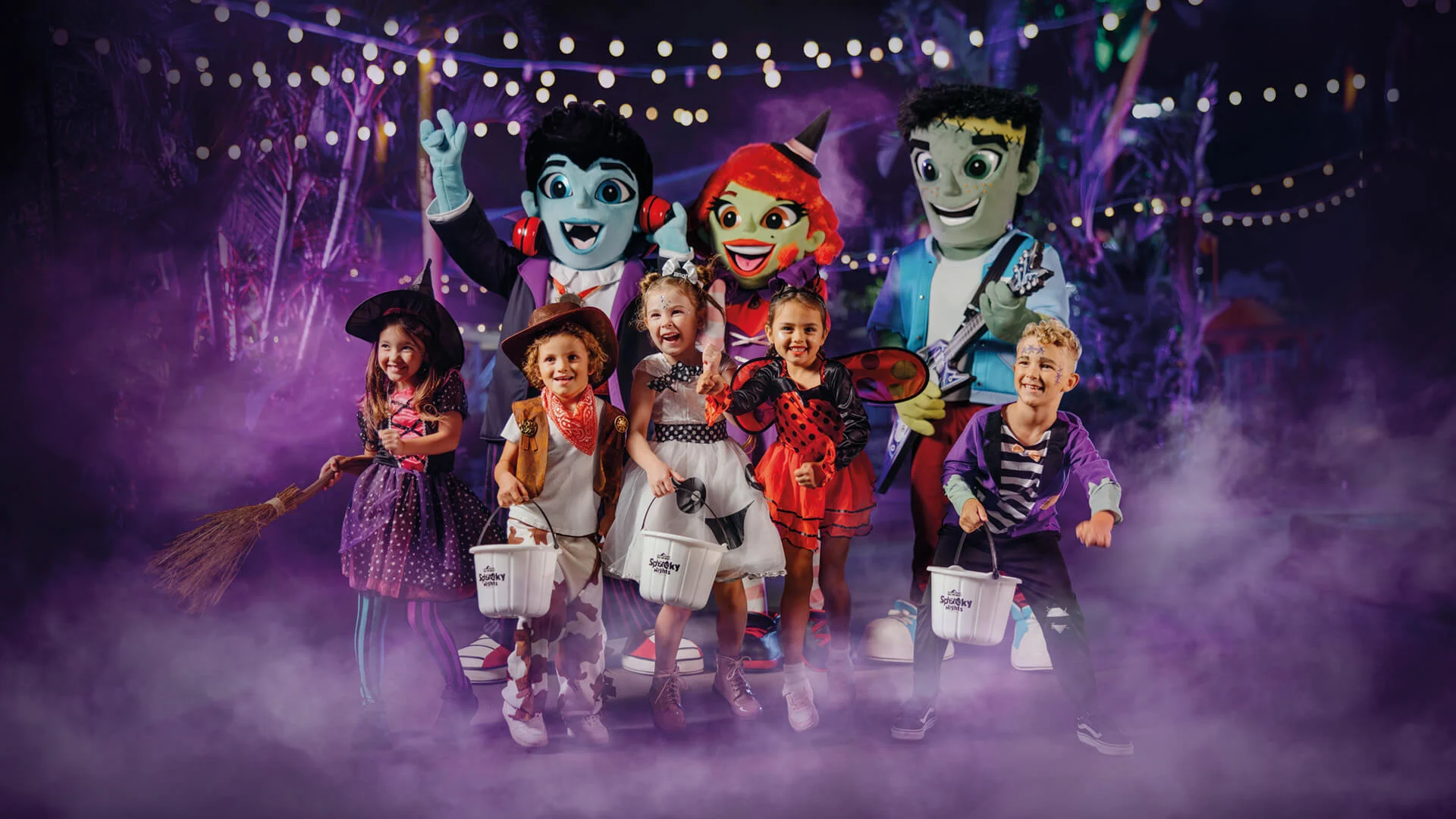 A group of young children in Halloween costumes stand smiling with trick-or-treat buckets, surrounded by friendly cartoon monster mascots, under string lights and purple mist in a festive nighttime setting.
