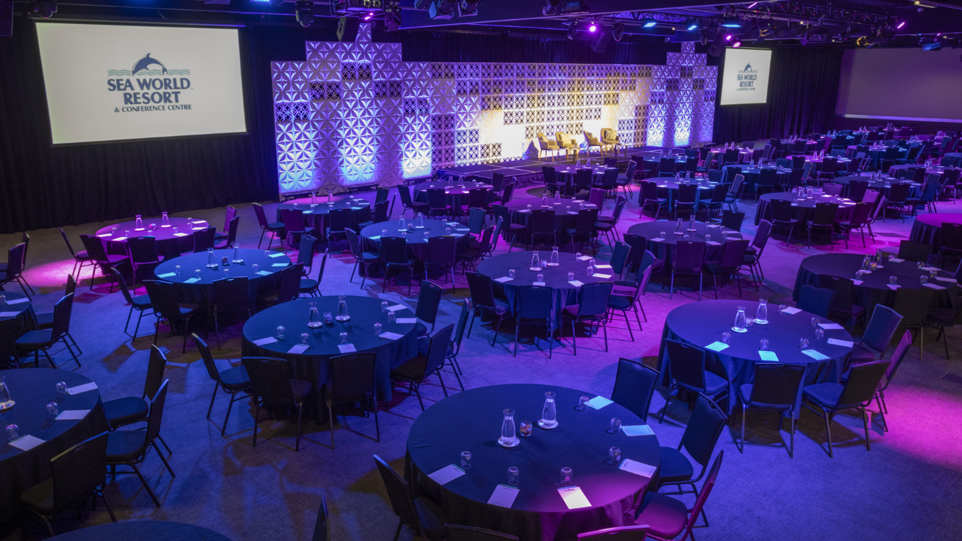 Conference room set up for an event with round tables, chairs, water bottles, and stage with screens displaying "Sea World Resort." Purple lighting highlights the space.