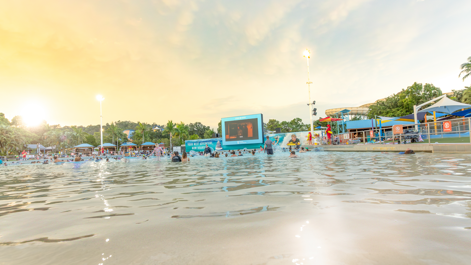 People are swimming and relaxing in a large wave pool at sunset, with a big screen and several buildings in the background, surrounded by palm trees and umbrellas. The sky has a warm, golden glow.