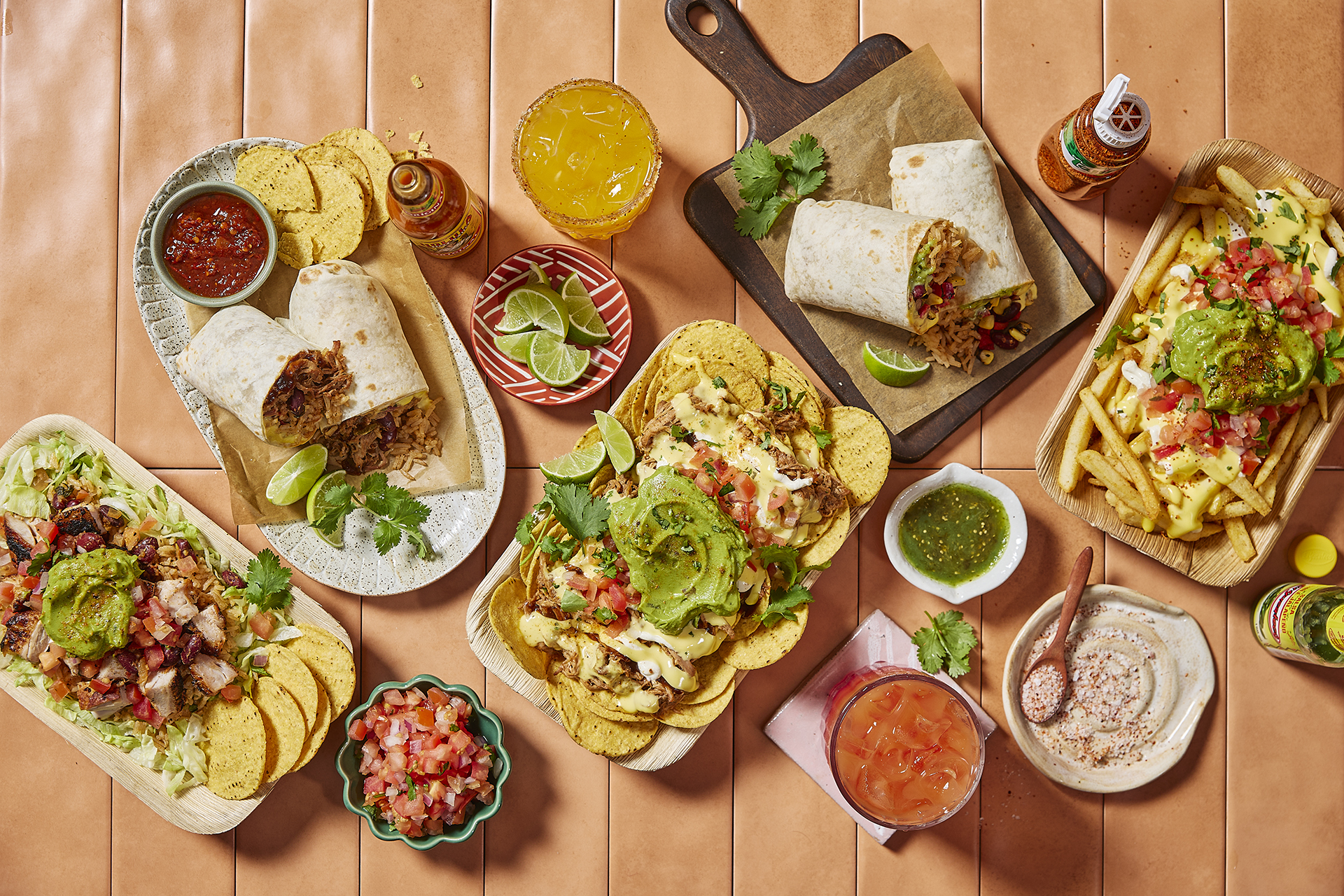 Assorted Mexican dishes including burritos, nachos, loaded fries, salsa, guacamole, and drinks arranged on a table with hot sauce bottles and lime wedges.