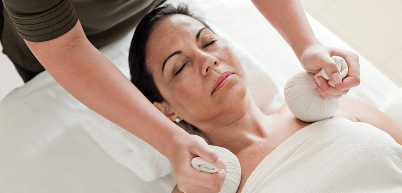 A person receiving a herbal compress massage on the neck and shoulders while lying on a massage table, covered with a towel.