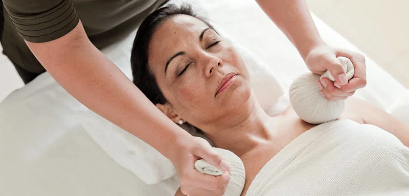 A person receiving a herbal compress massage on the neck and shoulders while lying on a massage table, covered with a towel.