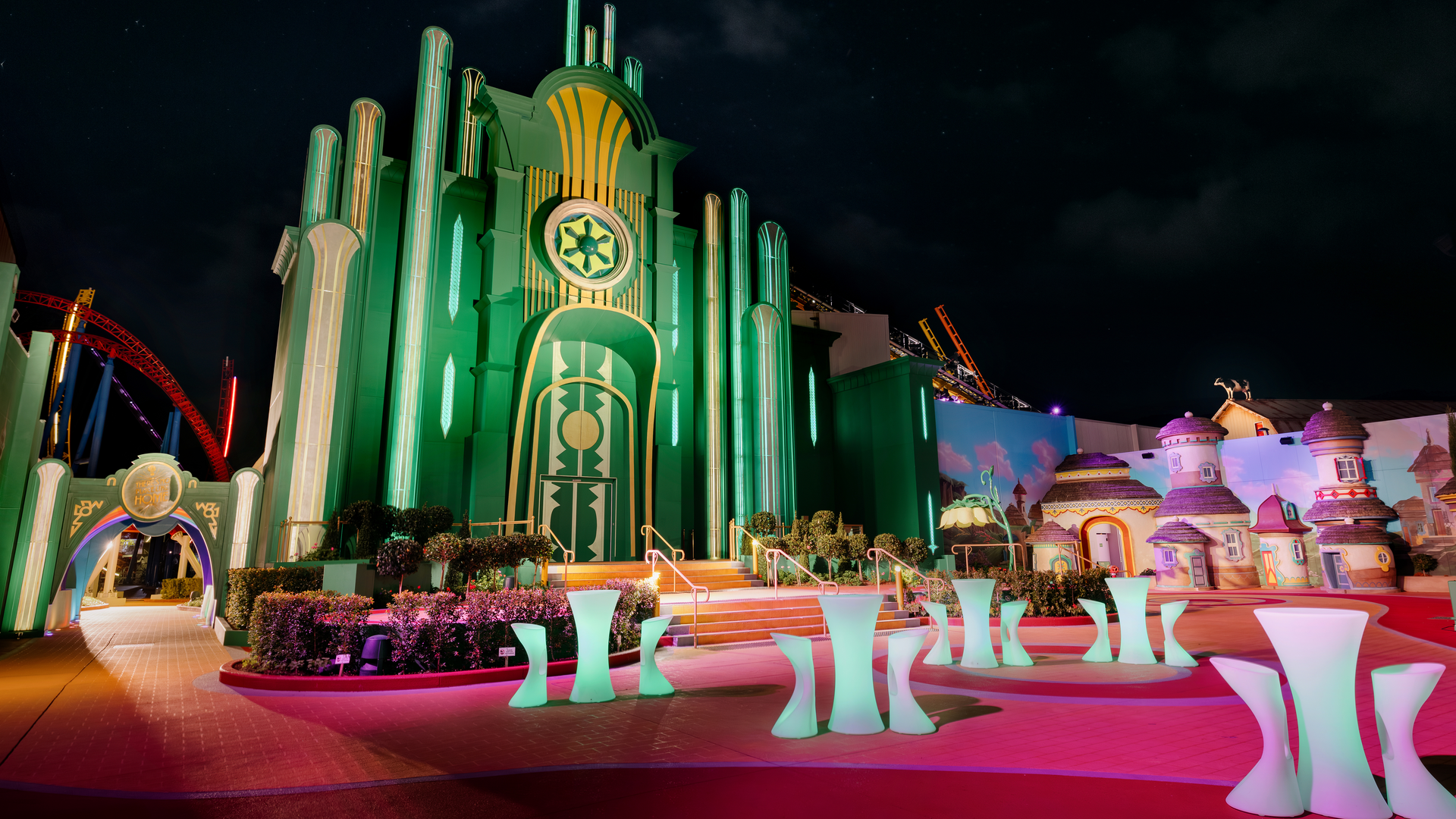 A brightly lit, green and yellow fantasy building with tall columns stands under a night sky, surrounded by colorful whimsical structures and glowing modern tables, resembling a scene from The Wizard of Oz.