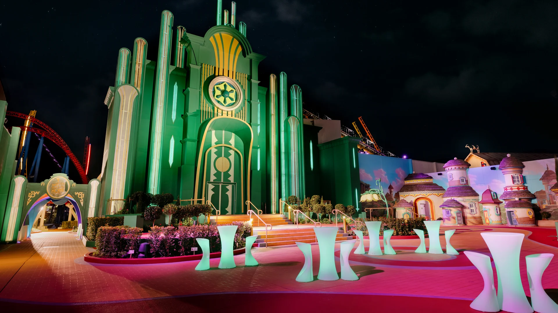 A brightly lit, green and yellow fantasy building with tall columns stands under a night sky, surrounded by colorful whimsical structures and glowing modern tables, resembling a scene from The Wizard of Oz.
