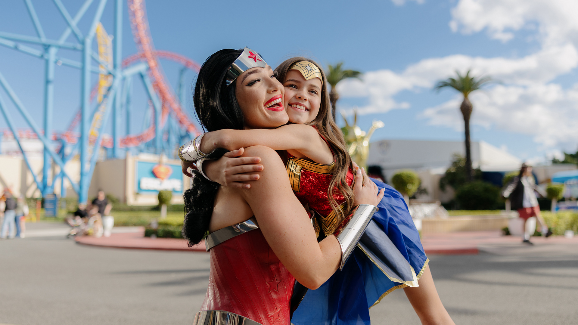 A woman and a young girl, both dressed as Wonder Woman, smile and embrace outdoors at an amusement park with roller coasters and palm trees in the background.