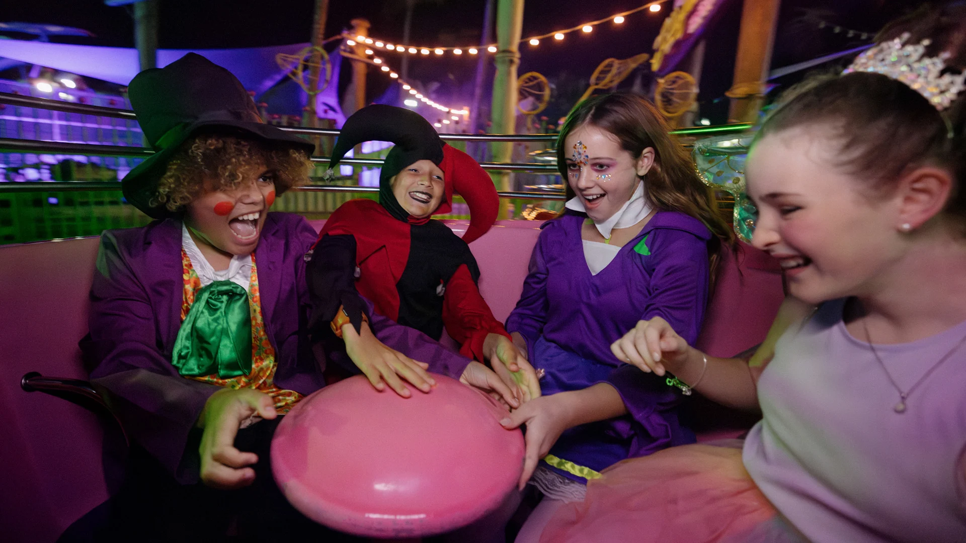 Four children in colorful costumes laugh and enjoy a spinning amusement park ride at night, sitting together in a pink seat with festive lights in the background.