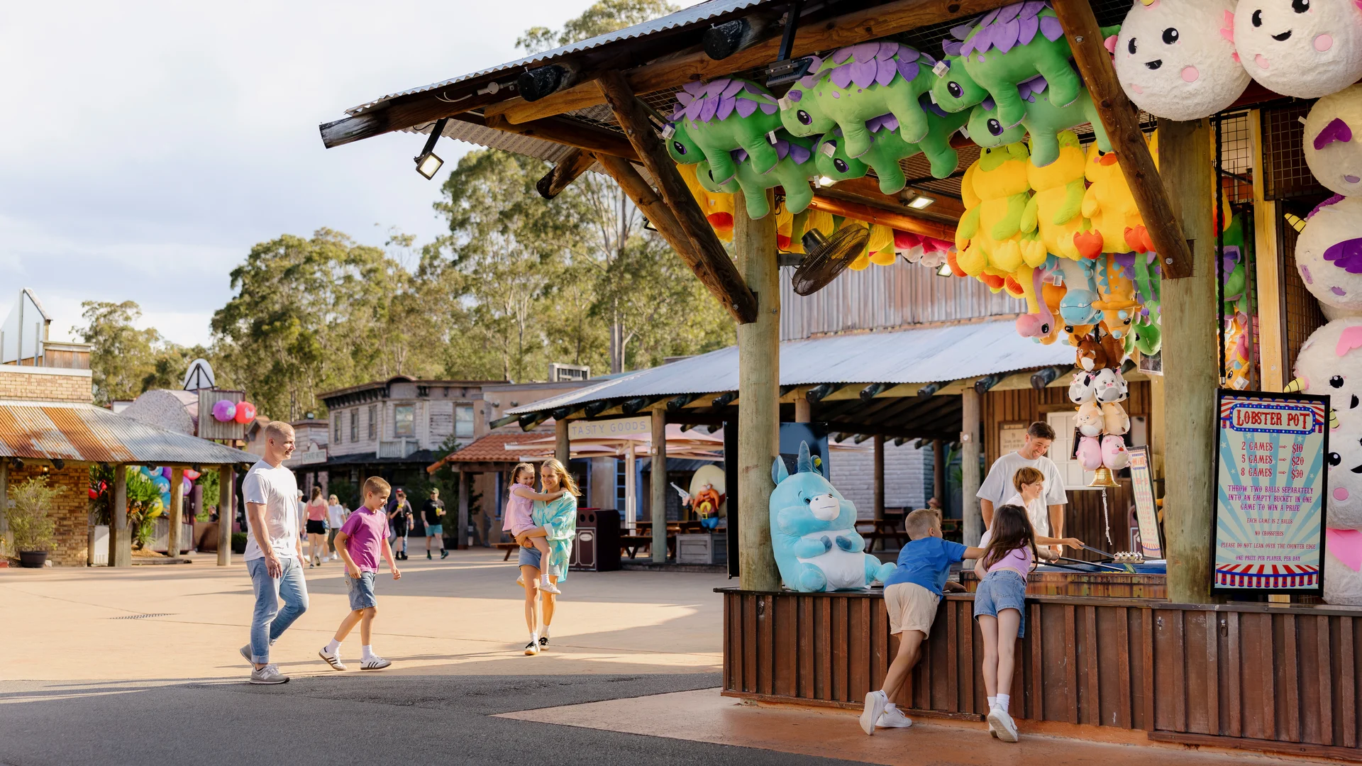 People enjoy a sunny day at an amusement park, walking past a colorful carnival game booth decorated with large plush toys, including animals and unicorns. Several children play the game while adults watch nearby.
