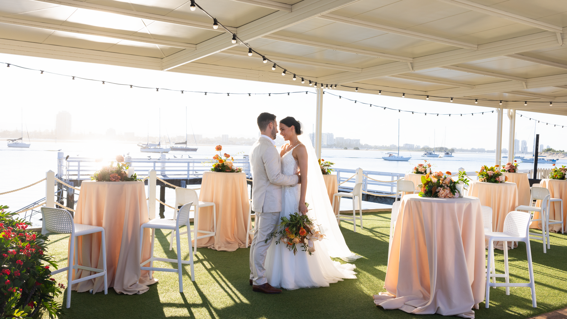A bride and groom stand close together under a covered, open-air venue decorated with string lights and pastel tablecloths, with boats and a city skyline visible in the background.