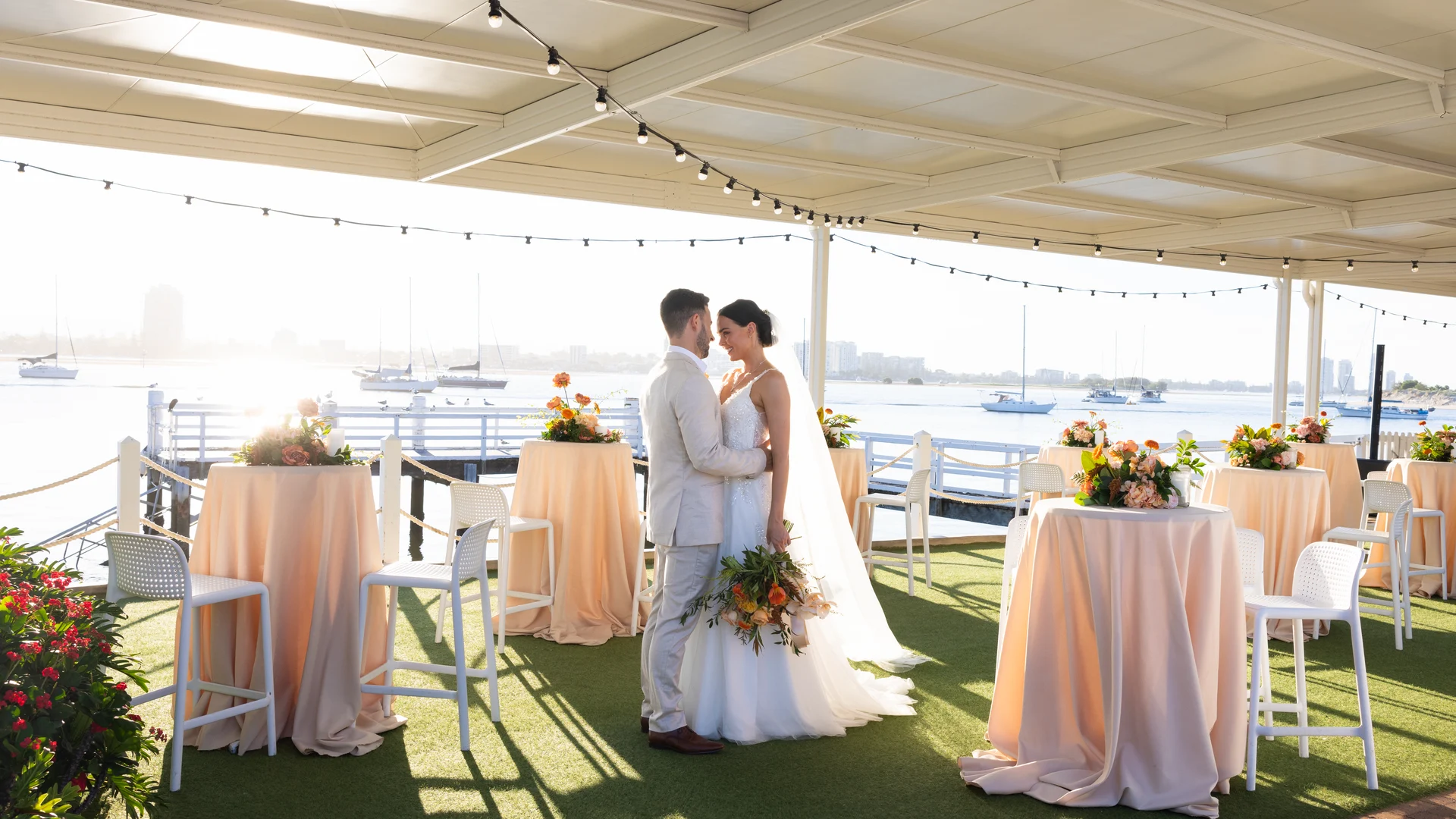 A bride and groom stand close together under a covered, open-air venue decorated with string lights and pastel tablecloths, with boats and a city skyline visible in the background.