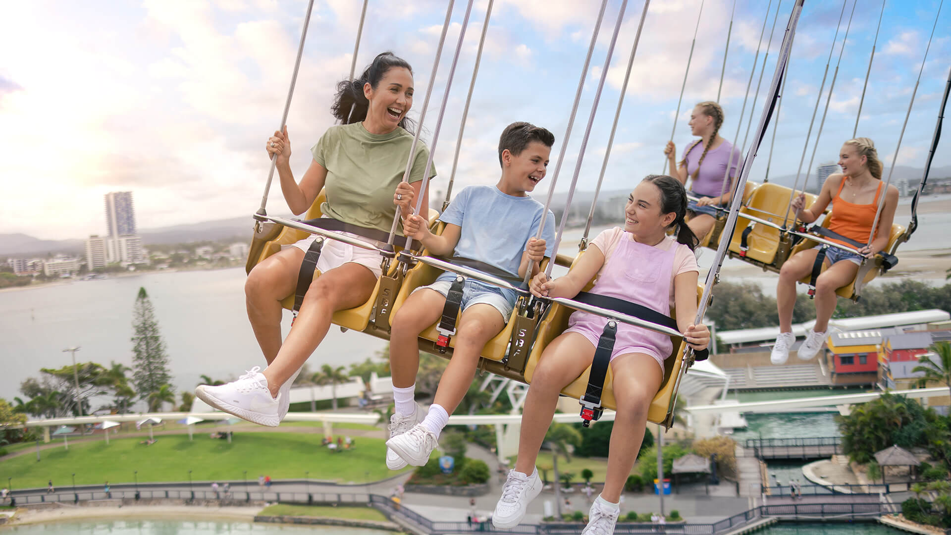 Three smiling teenagers ride a swing carousel at an amusement park, with water and buildings visible in the background under a partly cloudy sky. Other people are enjoying the ride as well.