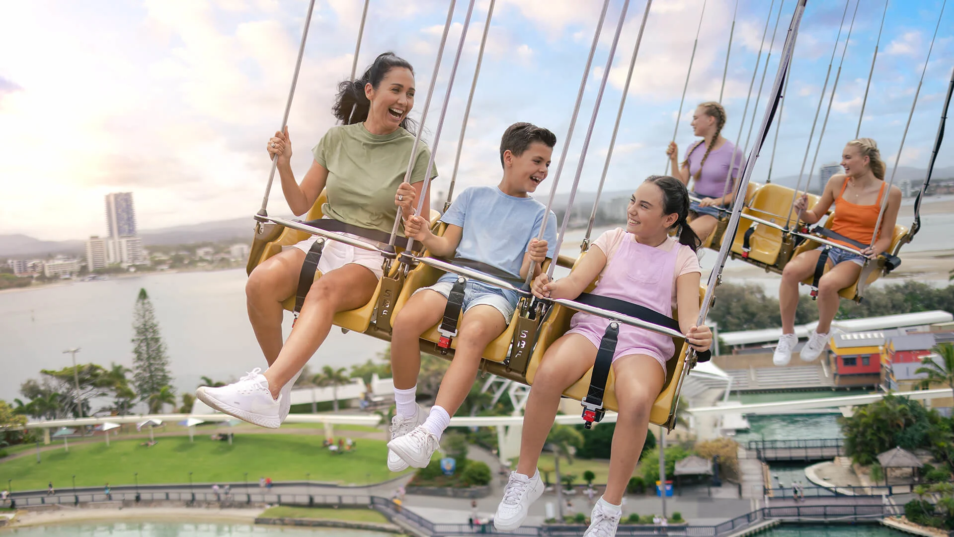 Three smiling teenagers ride a swing carousel at an amusement park, with water and buildings visible in the background under a partly cloudy sky. Other people are enjoying the ride as well.