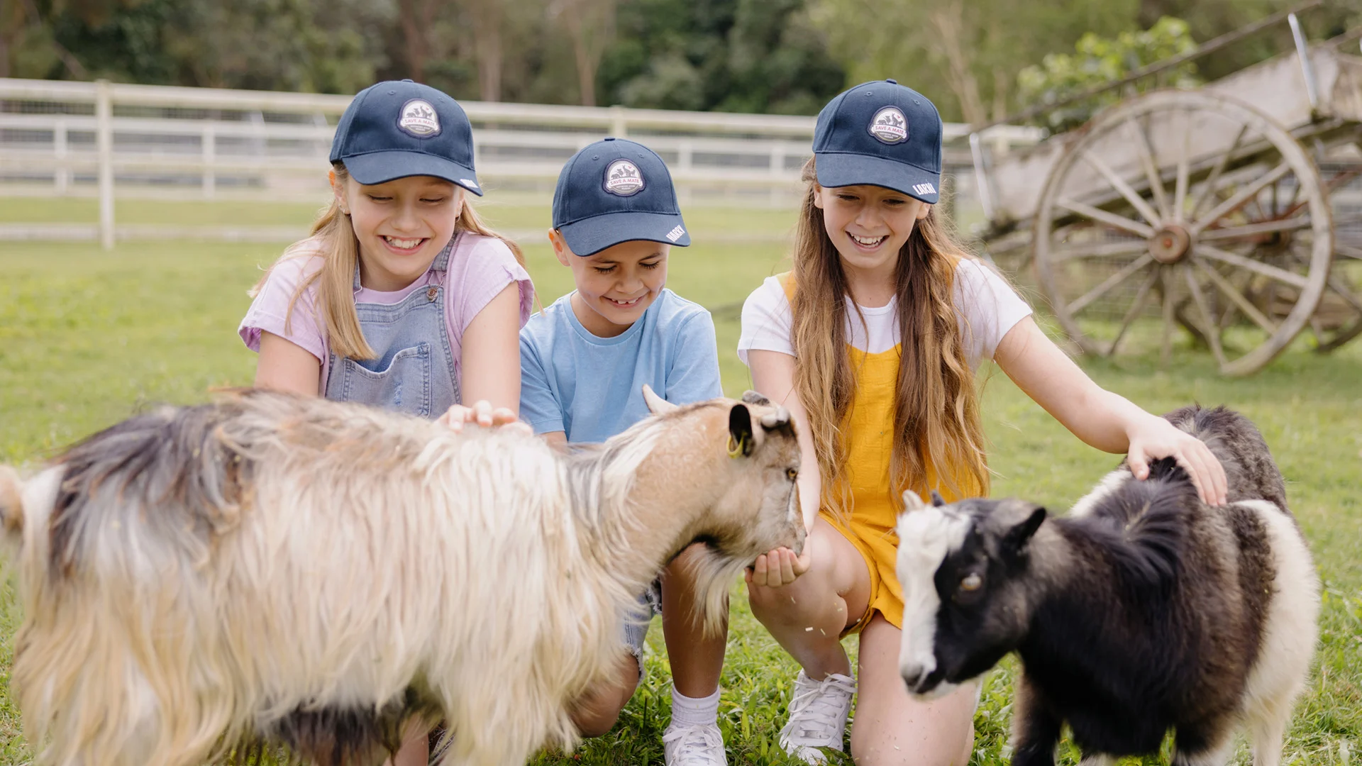 Three children wearing blue hats smile and pet two goats on a grassy farm, with a wooden wagon and fence in the background.