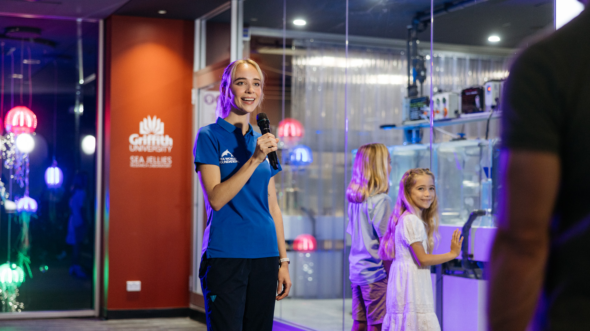 A woman in a blue shirt holds a microphone and speaks to visitors at a Griffith University Sea Jellies exhibit, with children nearby and colorful lights in the background.