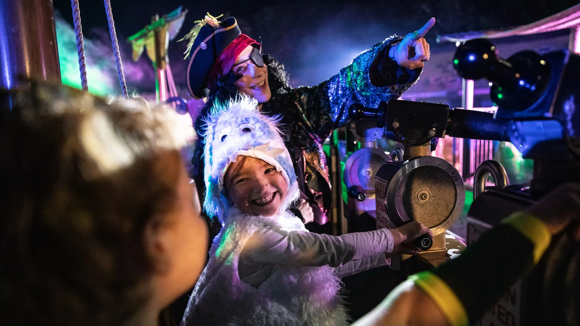 Two children in costumes, one dressed as a furry animal, smile and play with equipment under colorful lights while an adult in a pirate outfit points excitedly in the background. It appears to be a lively, festive event at night.