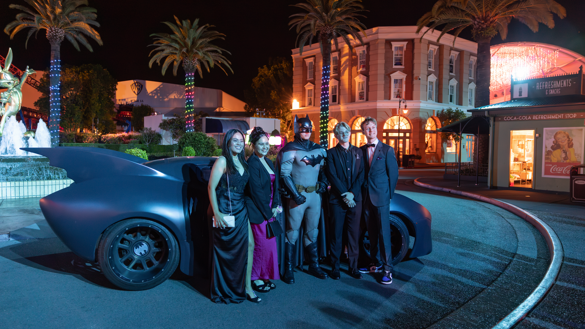 A group of five people dressed formally and one in a Batman costume pose in front of a Batmobile replica at night, surrounded by palm trees and illuminated buildings.