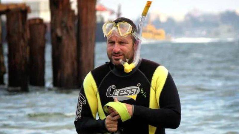 A man in a wetsuit and snorkel stands near the shore, holding yellow swim fins, with wooden pilings and water in the background.