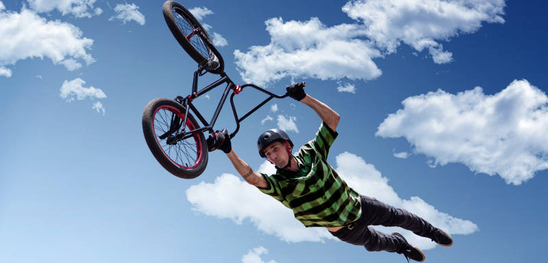 A person wearing a helmet and striped shirt performs an aerial trick on a BMX bike against a blue sky with scattered clouds.