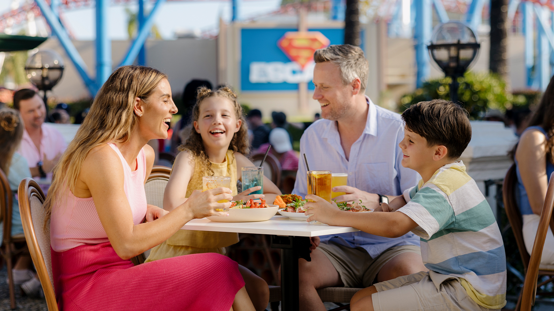 A smiling family of four sits at an outdoor table, enjoying food and drinks at an amusement park on a sunny day. Roller coaster tracks and a Superman sign are visible in the background.