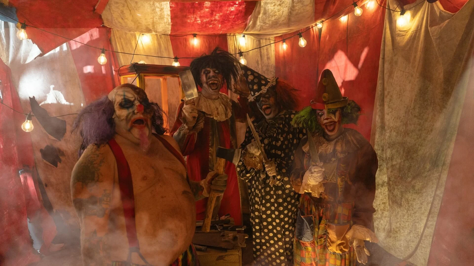 Four people dressed as sinister, scary clowns stand under string lights in a red-and-white circus tent, surrounded by fog. They wear exaggerated makeup and costumes, with one clown holding an axe and another in a polka-dot suit.
