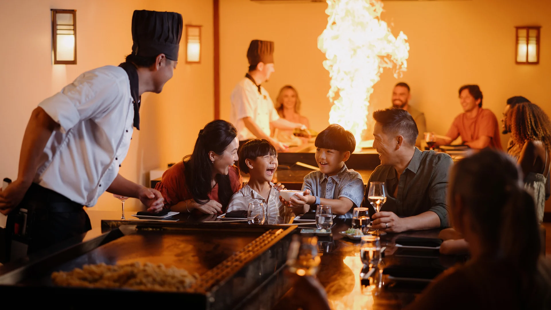 A chef in a black hat entertains a smiling family with a fiery teppanyaki grill show at a restaurant, while other diners watch and laugh in the warmly lit background.