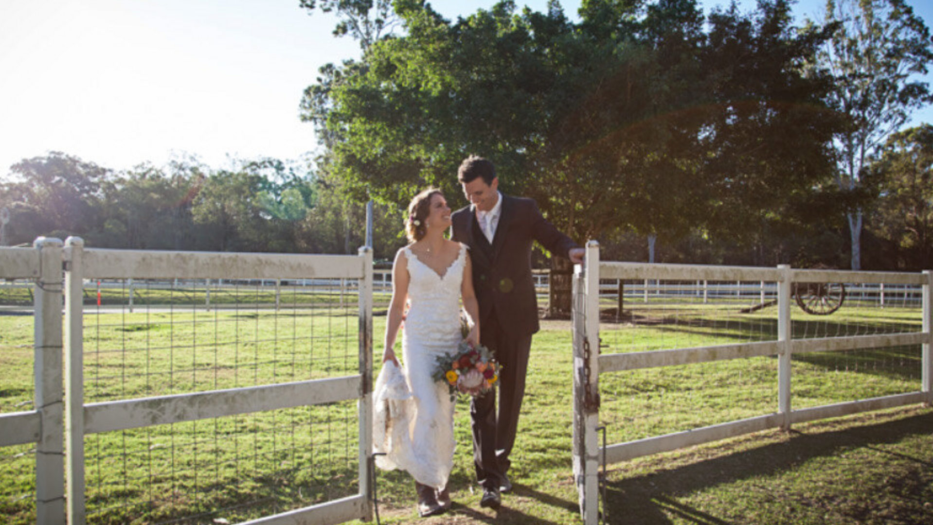A bride and groom walk together through an open gate on a sunny day in a grassy, fenced outdoor setting.
