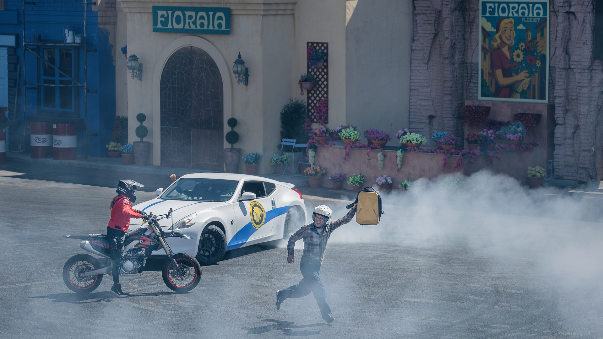 A person in a helmet runs holding a gas can while a motorcyclist and a white car create tire smoke in front of a Floralia-themed building decorated with flowers.