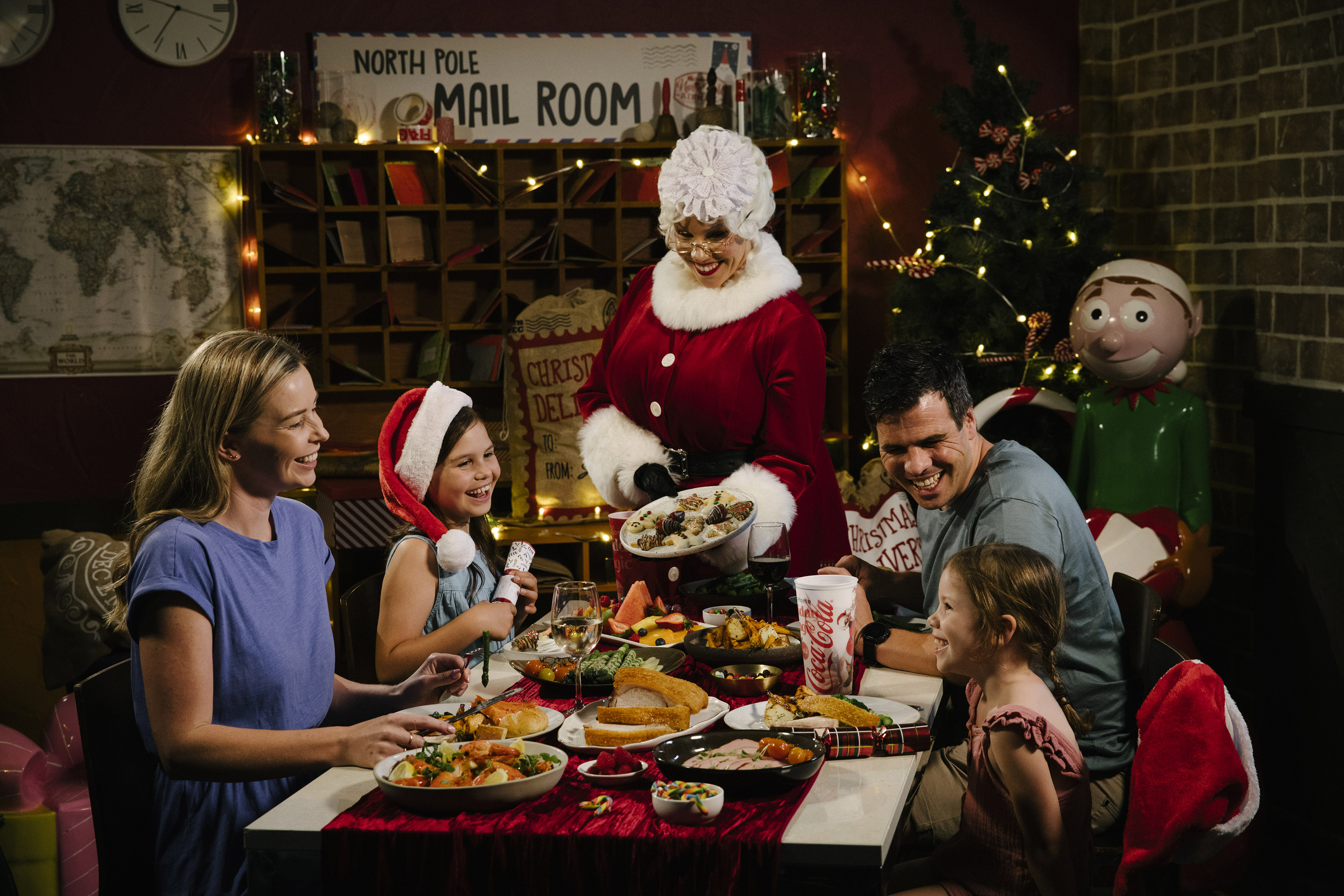 A family of four smiles at a festive dinner table as a woman dressed as Mrs. Claus serves cookies. The setting is decorated for Christmas, with a tree, holiday lights, and a "North Pole Mail Room" sign.