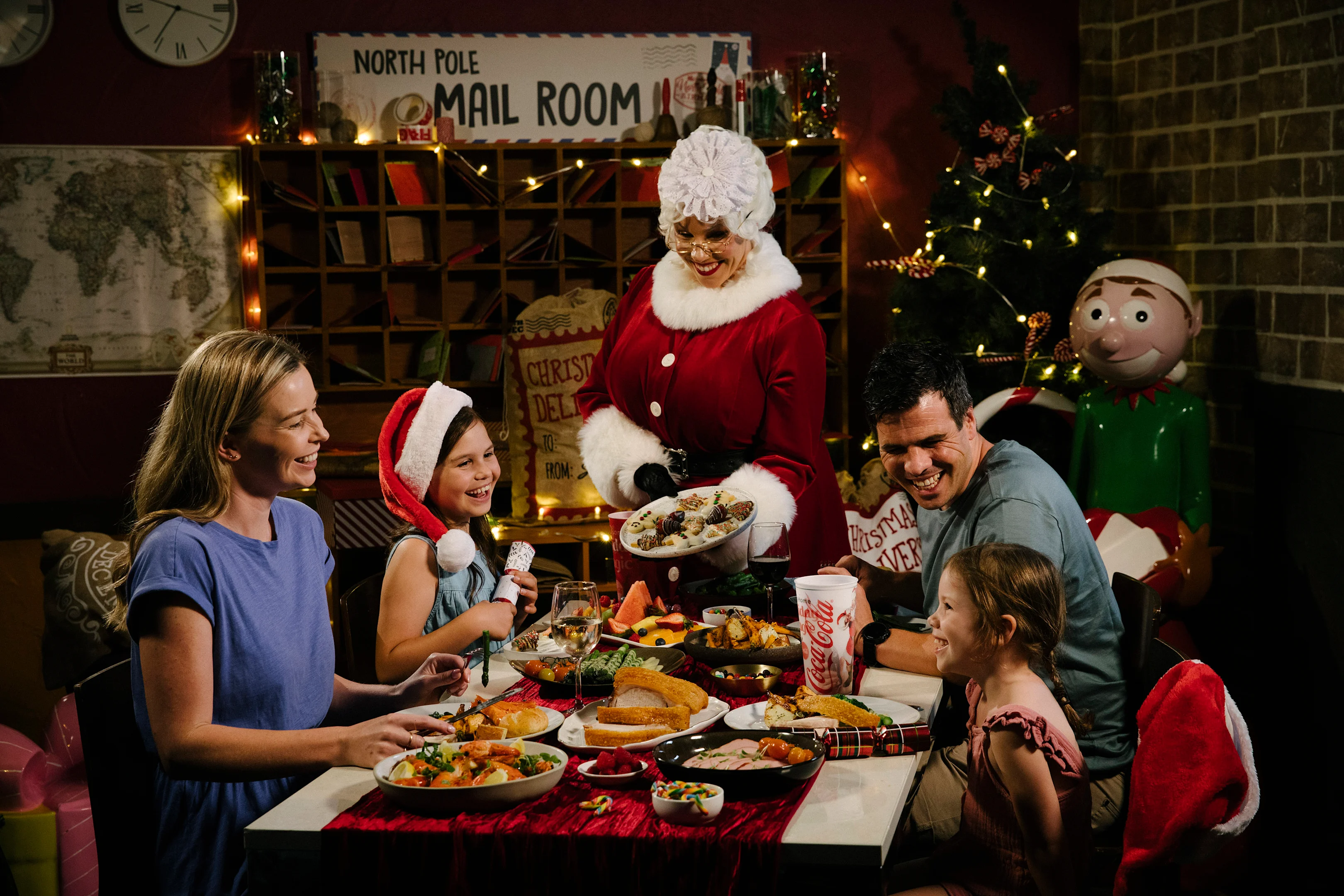A family of four smiles at a festive dinner table as a woman dressed as Mrs. Claus serves cookies. The setting is decorated for Christmas, with a tree, holiday lights, and a "North Pole Mail Room" sign.