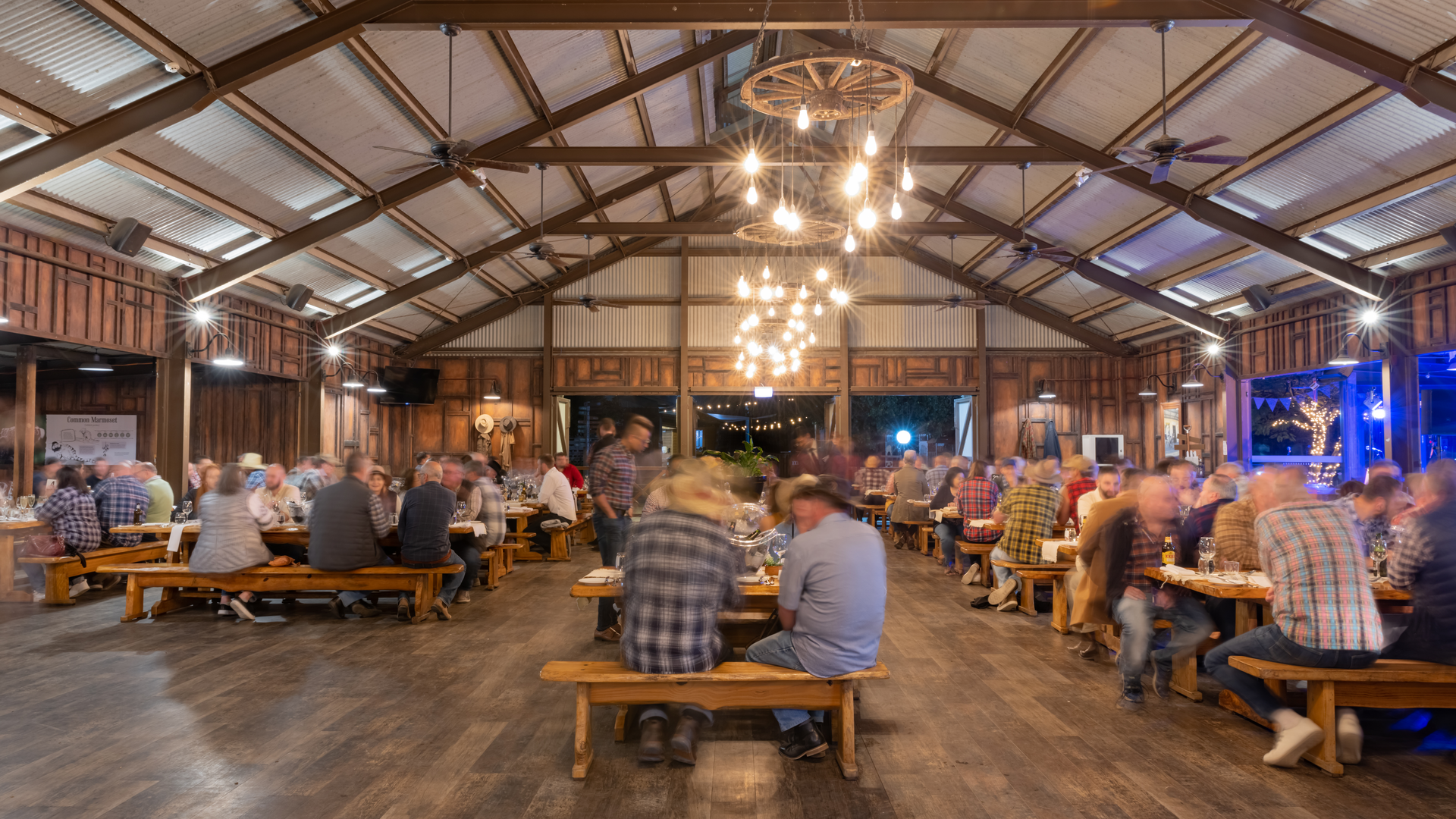 People are seated at wooden tables inside a large, rustic hall with exposed beams and hanging lights, creating a lively dining atmosphere.