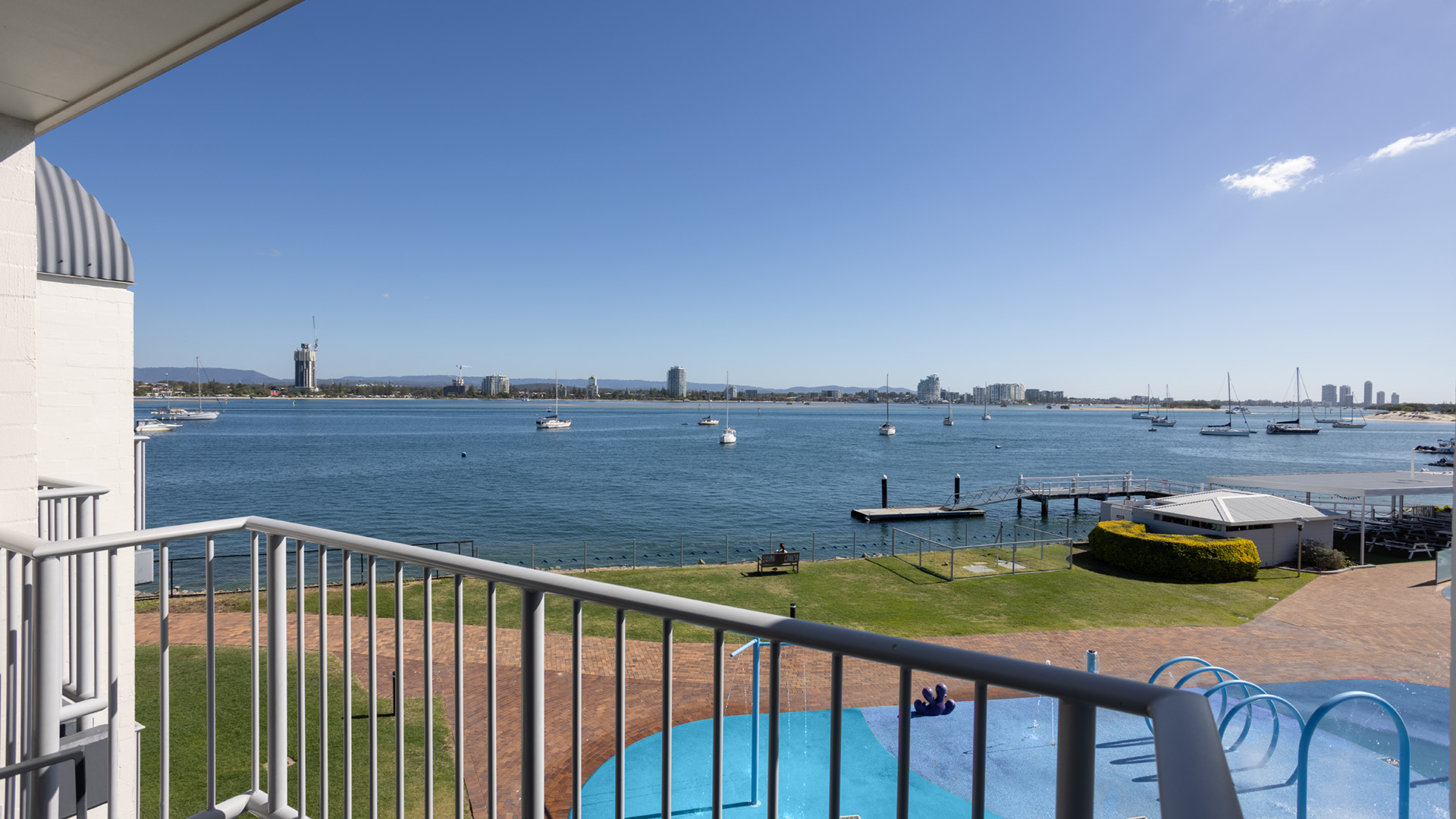 View from a balcony overlooking a large body of water with several boats and distant city skyline. A railing and a small park area with pathways and a children's play area are visible.