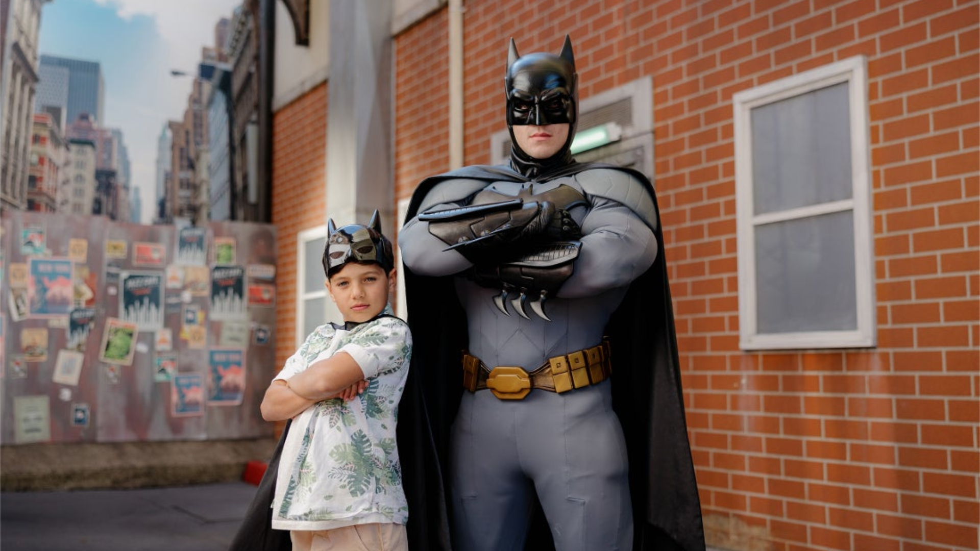 A child and an adult, both wearing Batman masks and capes, stand confidently with arms crossed in front of a brick building in a city setting. The adult is dressed in a full Batman costume.