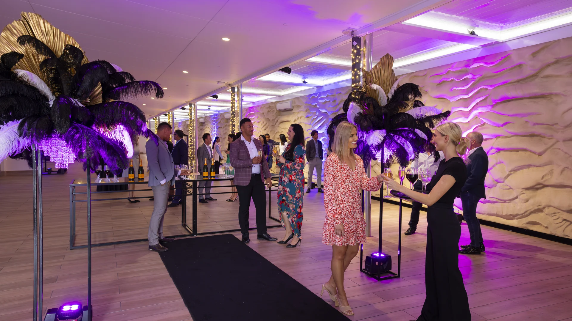 People mingling at a formal event with purple lighting, ornate palm decorations, and a table with champagne bottles.
