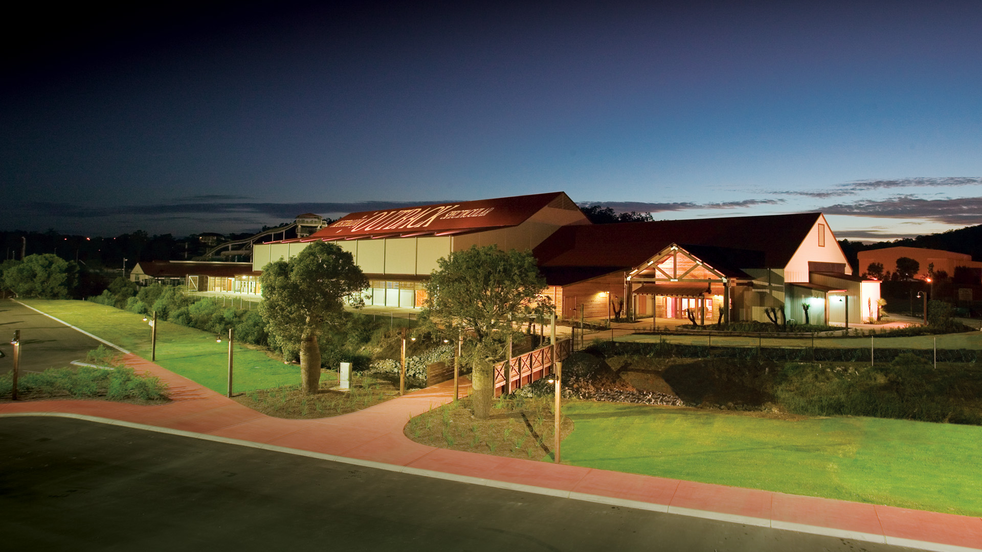 A large, modern building with a red roof and illuminated exterior stands surrounded by green lawns and trees at dusk, with a clear sky and soft outdoor lighting creating a welcoming atmosphere.