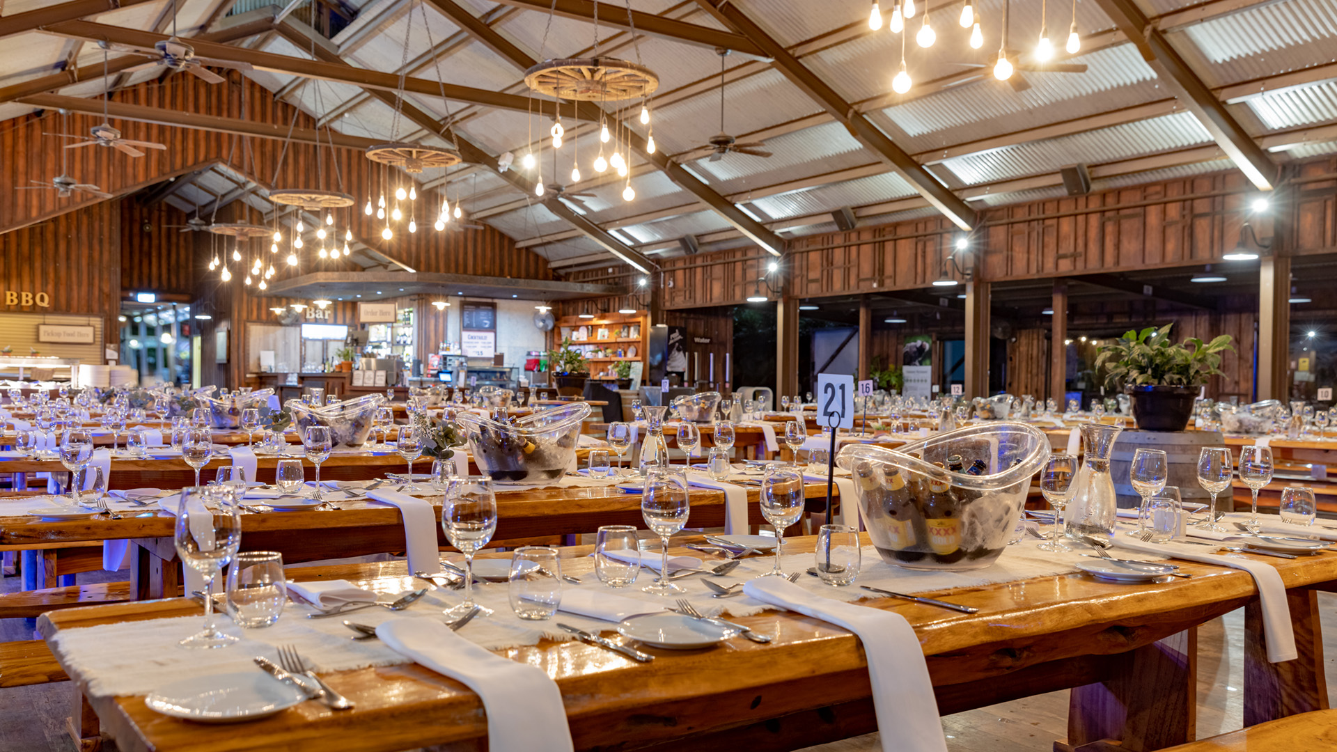 A spacious, rustic banquet hall with long wooden tables set for an event, featuring wine glasses, cutlery, and napkins. Warm hanging lights illuminate the room, giving it a cozy and inviting atmosphere.
